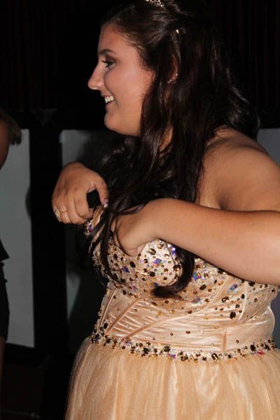 Woman in a peach-colored formal dress adjusts the bodice, smiling. Dark hair, tiara, and a dance setting.