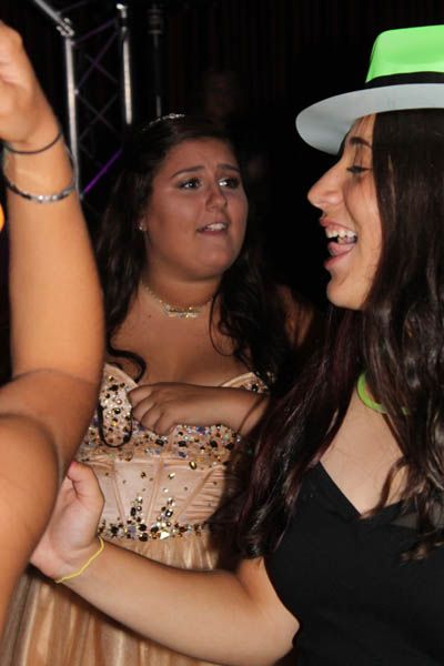 Two young people dancing at a party; one wearing a hat and smiling, the other looking surprised in a formal gown.