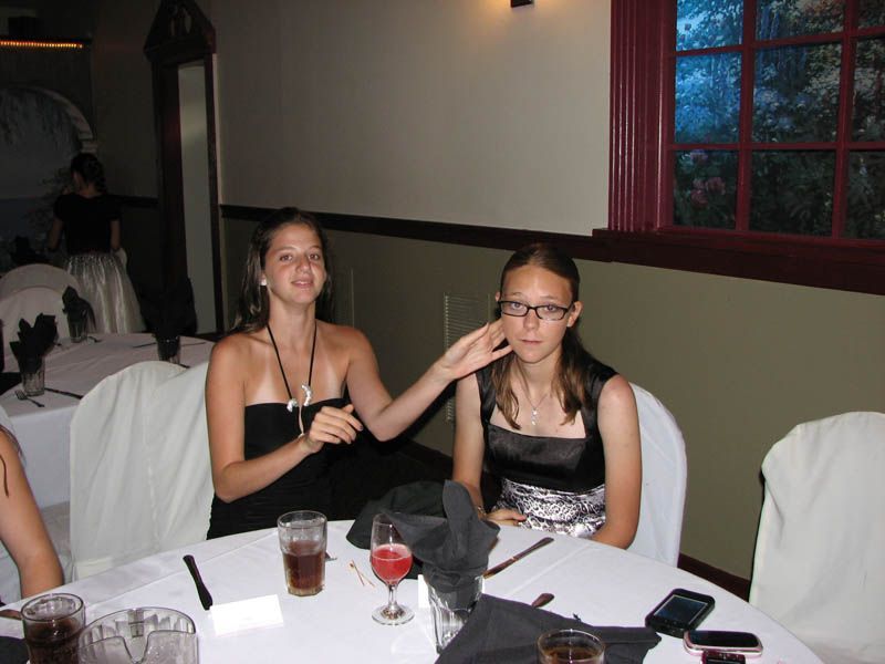 Two women at a table; one touches the other's face. Both in formal dresses. Indoor setting with drinks.