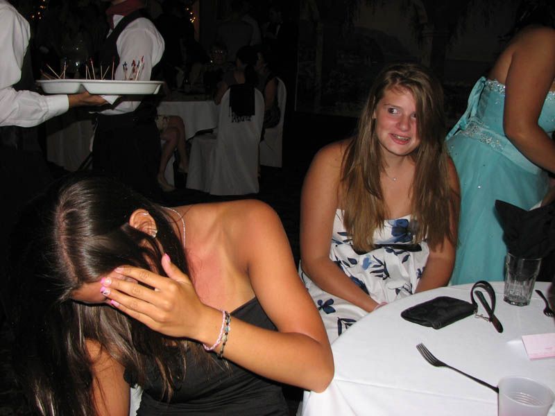 Two young women at a formal event, one covering her face, the other looking surprised. Server with drinks.