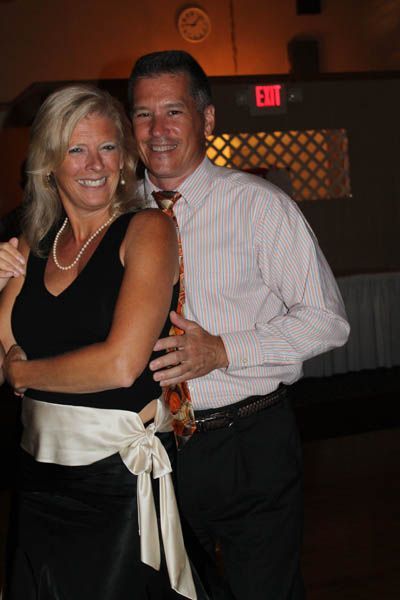 Couple dancing close together at an event. Man in a button-down shirt, woman in a black dress with a cream sash.