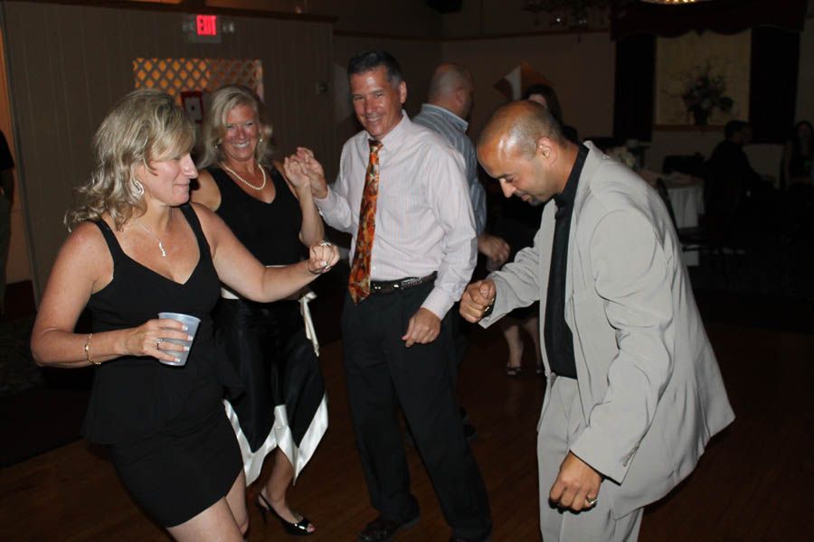 People dancing at a gathering. Woman in black dress holds a cup, others smile and dance. Indoors.