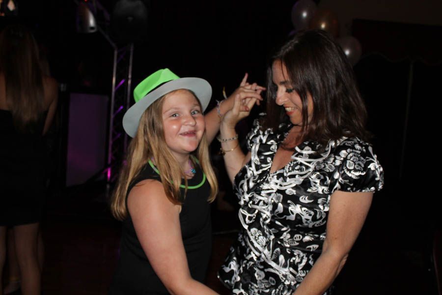 Woman and child dancing in a dark room; child wears a green hat and glow necklace, smiling.