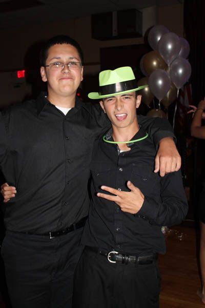 Two young men in black shirts at an event. One wears glasses and smiles, other makes peace sign and has a green hat.