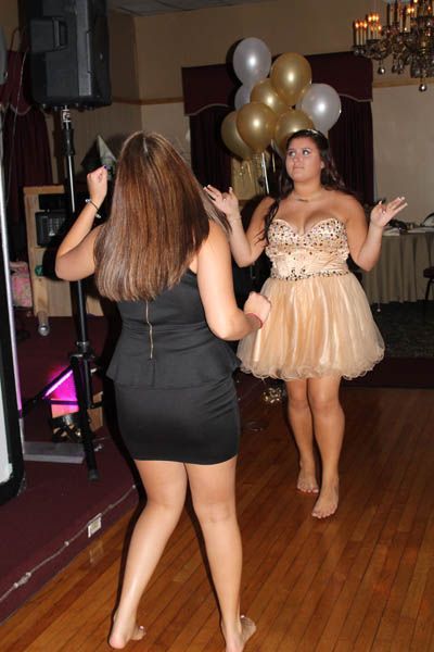 Two women dancing barefoot in a room with balloons. One wears a gold dress, the other a black dress.