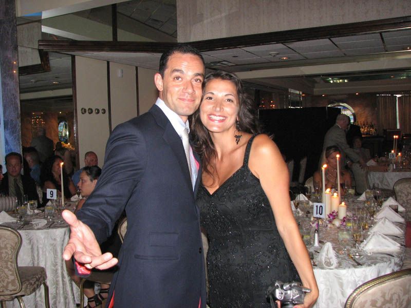 Couple smiles, posing at a formal event, near decorated tables. The man wears a suit, and the woman a black dress.