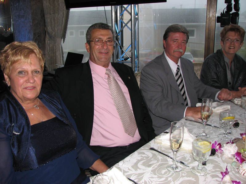 Four people sit at a decorated table, dressed formally. Two men wear suits and ties, two women wear formal tops.