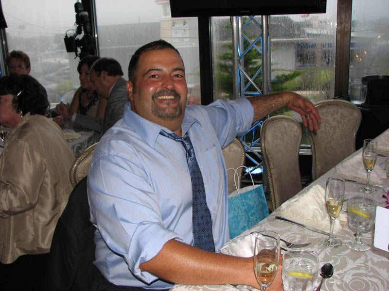 Man in blue shirt, tie, seated at a formal table, smiling; restaurant interior with others present.
