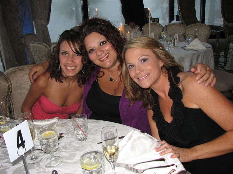 Three women at a table smiling, one with arm around others. Formal event with table settings and a number 