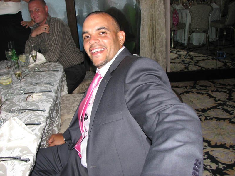 Man in suit smiles, seated at a decorated table, pink tie. Another man in background.