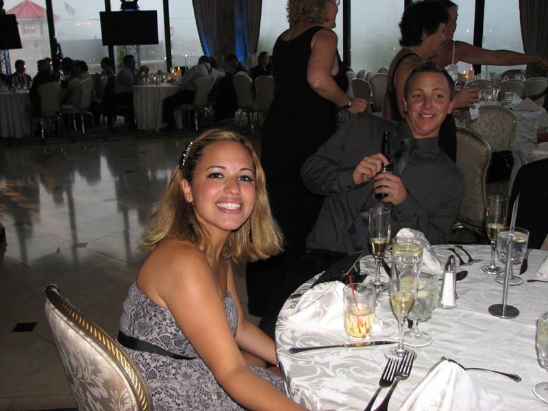 Woman in strapless dress smiles at a table; man in gray shirt laughs. Formal event setting with other guests.