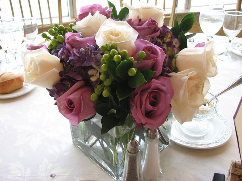 Arrangement of white and purple roses, purple hydrangeas, and green berries in a square glass vase on a table.