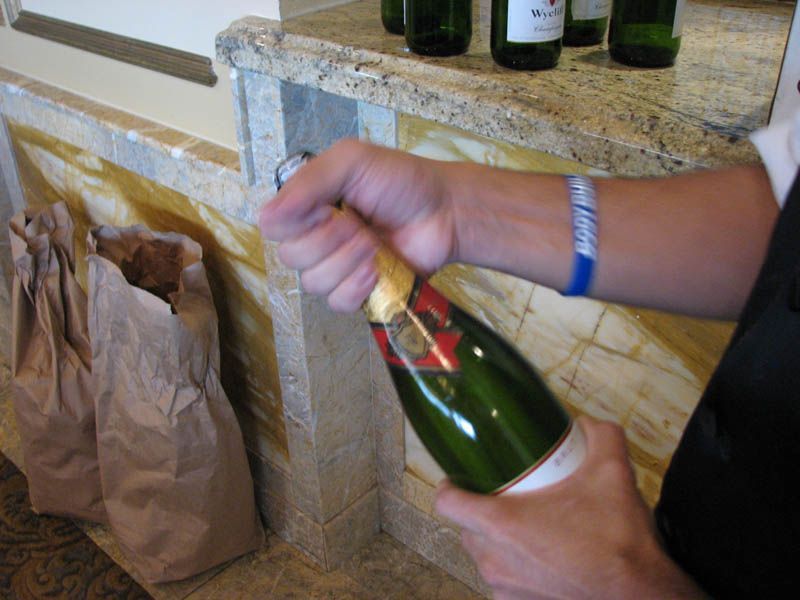 Person opening a green champagne bottle near a stone counter with other bottles and brown paper bags.
