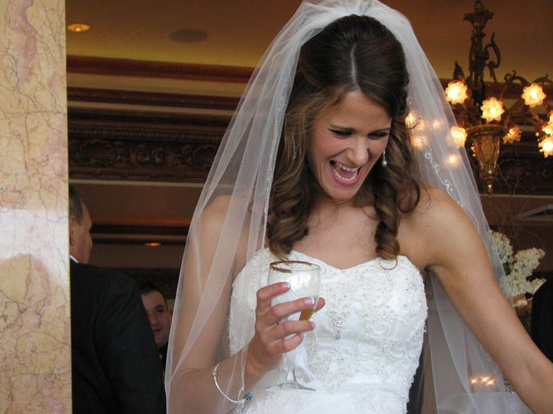 Bride in a white gown laughing, holding a drink, veil, with guests and chandelier in the background.