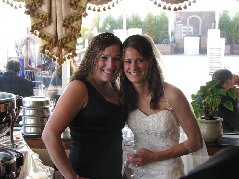 Bride in white dress smiles with woman in black dress; wedding reception.