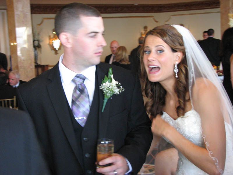 Bride in wedding dress and groom in suit, looking at each other, bride open-mouthed, room with guests.