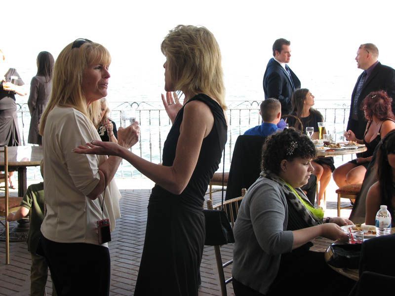 Two women conversing at an event, gesturing with hands; other guests in the background. Bright setting.