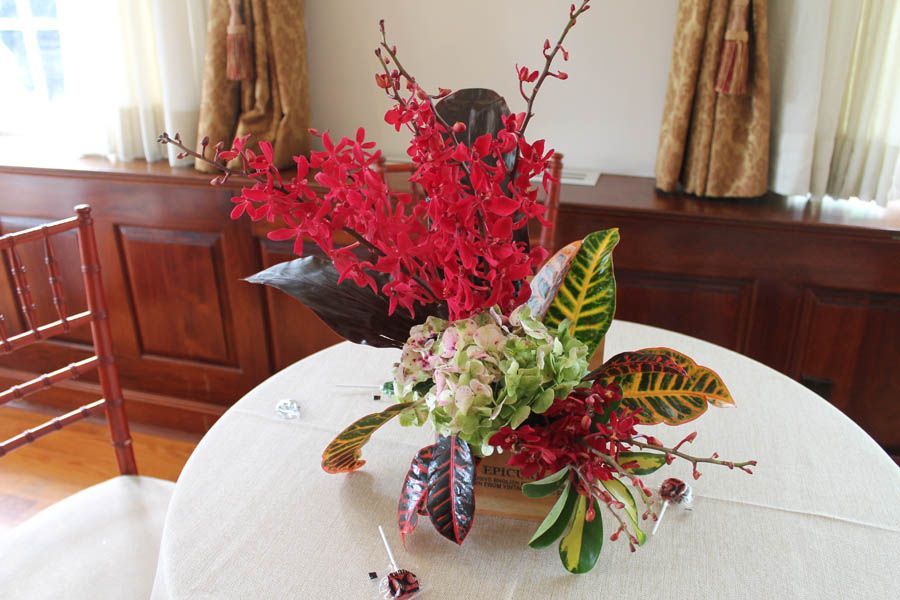 Flower arrangement on a round table, featuring red and green flowers and foliage, set in a room with wood paneling.