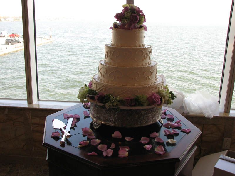 Five-tiered wedding cake with floral decorations on a table, petals and water in background.