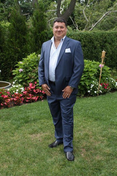 Man in blue suit, white open shirt, standing on grass in garden setting.