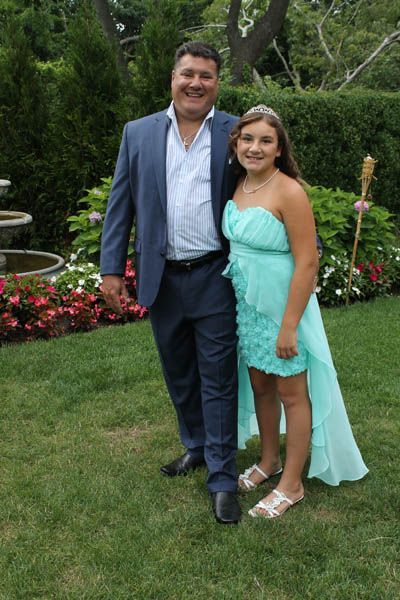 Man in a blue suit stands with girl in a teal dress, a tiara, and sandals on a lawn.