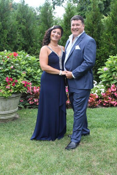 Couple in formal attire holding hands, smiling. Standing in a garden with flower beds and trees.