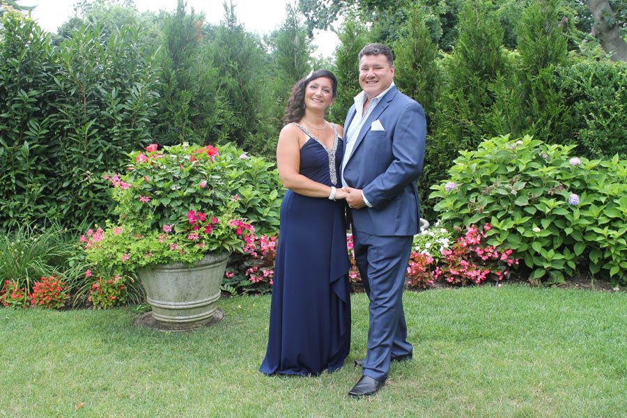 Couple in formal attire, holding hands, pose in garden with flowers and bushes.