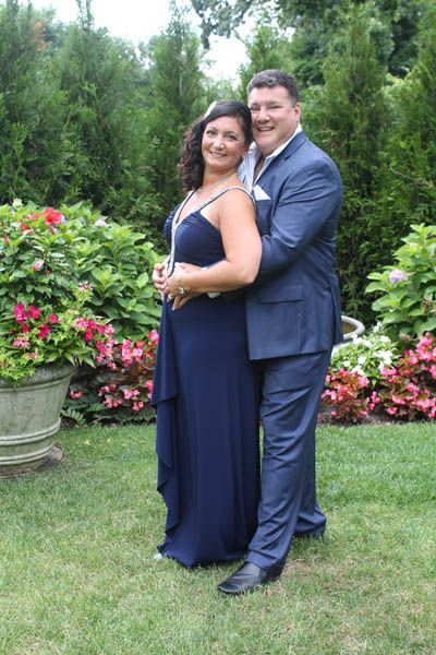 Couple embraces outdoors. Woman in blue gown, man in blue suit, smiling. Surrounded by greenery.
