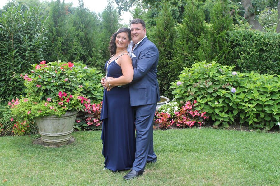 Couple in formal attire embracing in a garden, greenery and flowerpots in background.