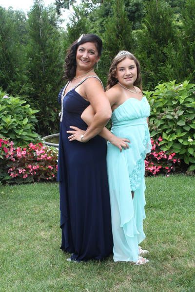Woman and girl in formal gowns stand back-to-back in a garden.