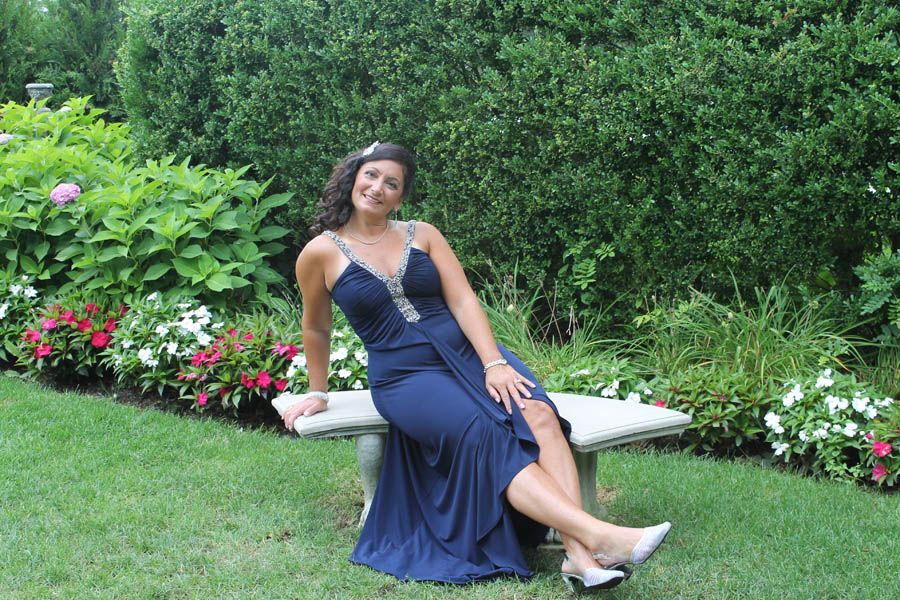 Woman in navy gown sits on bench in garden, smiling.