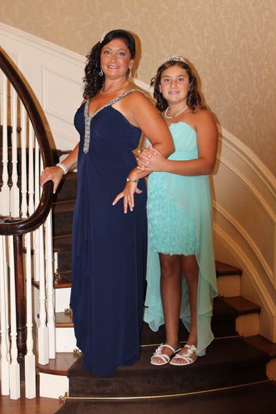 Woman in navy gown stands beside girl in teal dress on staircase.