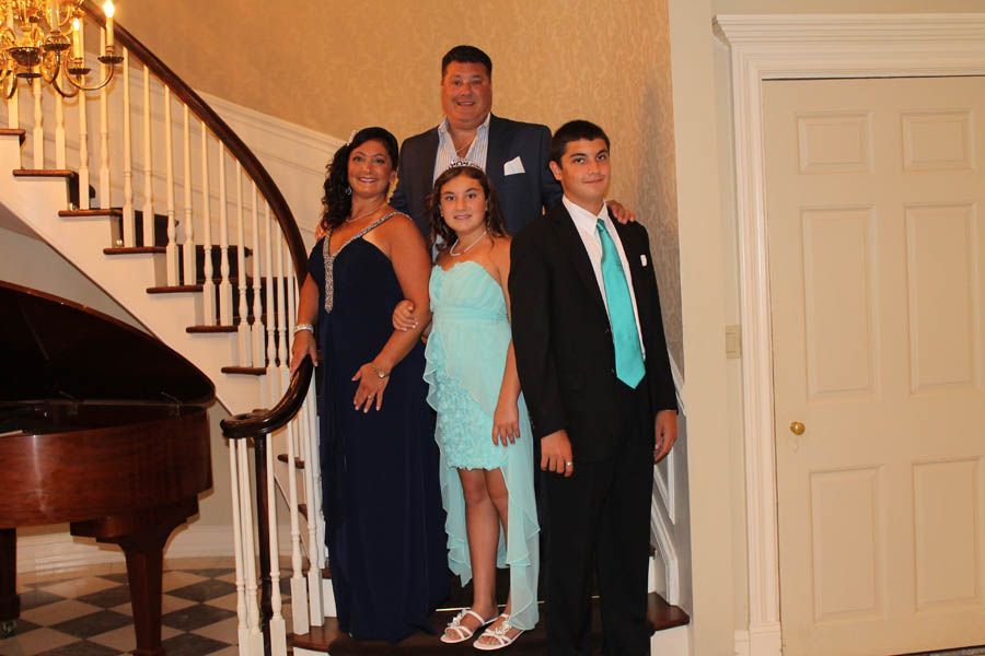 Family posing on a staircase, formal attire. Woman in blue, girl in aqua dress, man in suit. Piano and door in background.