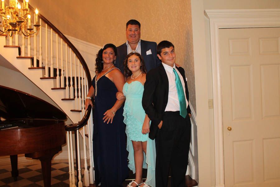 Family of five posing on a staircase, formal attire. Woman in blue gown, girl in turquoise dress, man in suit. Piano visible.
