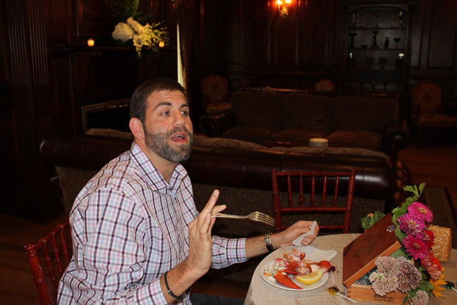 Man gesturing while seated at a table with food, in a dark wood-paneled room.