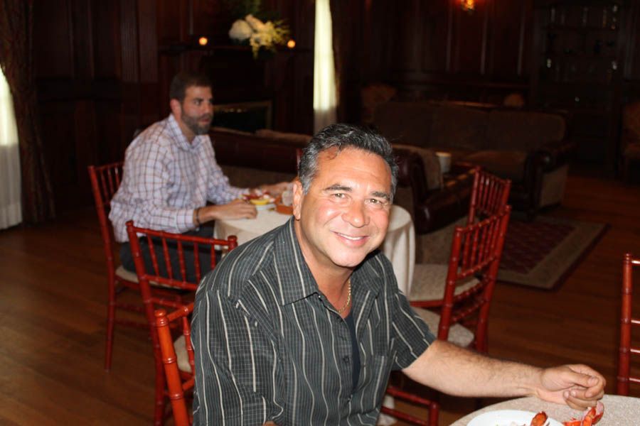 Man smiles at the camera at a table; another man sits in the background. Interior of a room.