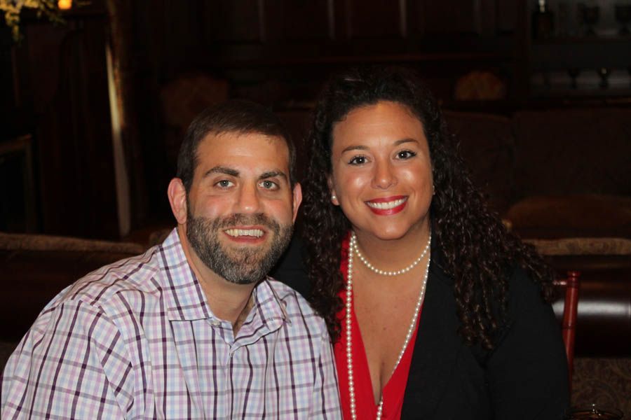 Man and woman smiling, sitting close together. Man in plaid shirt, woman in black top with pearl necklace.