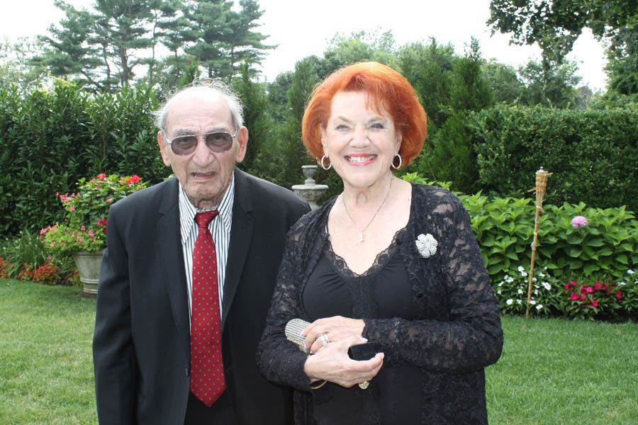 Elderly man and woman smiling outdoors, man in suit, woman in black lace, green background.