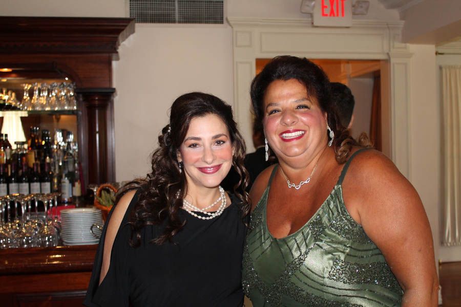 Two smiling women posing together indoors, near a bar. One wears a black dress, the other a green beaded gown.