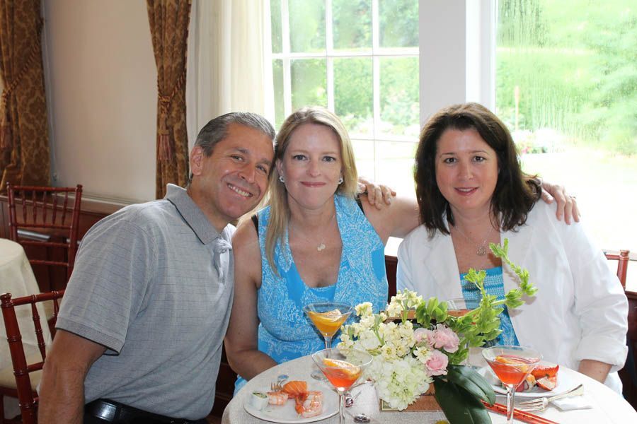 Three people smiling, sitting at a table with drinks and flowers near a window.