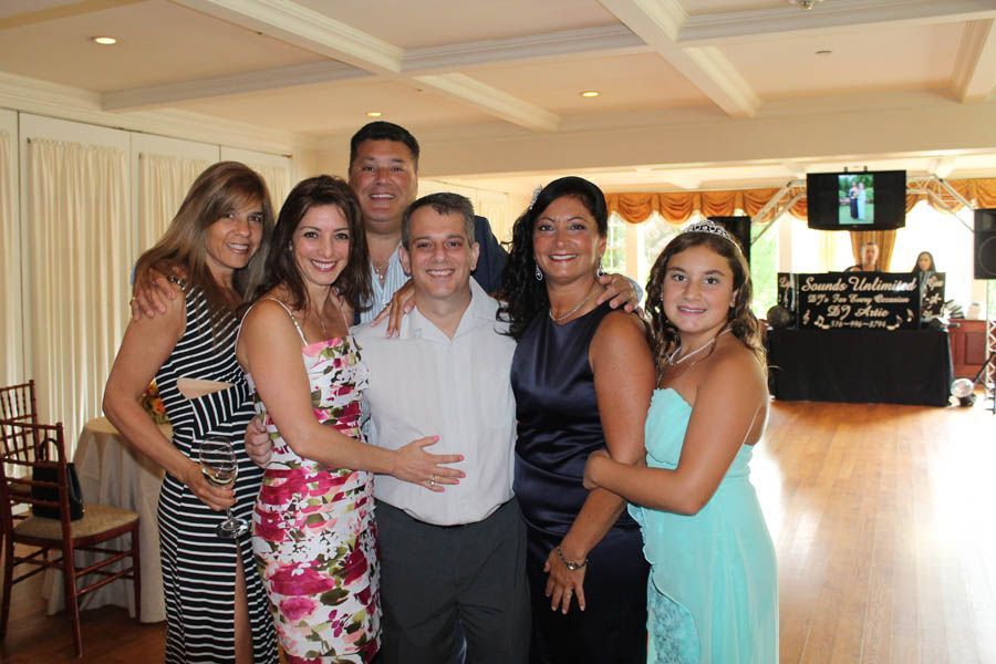 Group of six people smiling, posing together at an event. Indoors, with a dance floor and DJ setup in the background.
