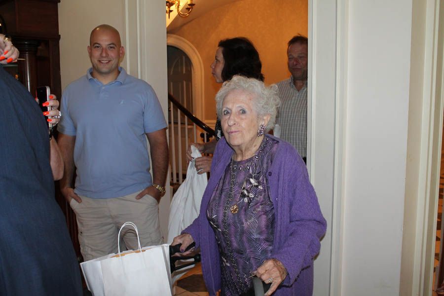 Elderly woman in purple sweater with a cane, entering a building, with others nearby.