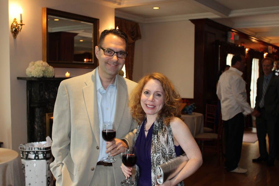 Man and woman at an event, holding wine glasses, smiling. Interior setting with fireplace and other guests.