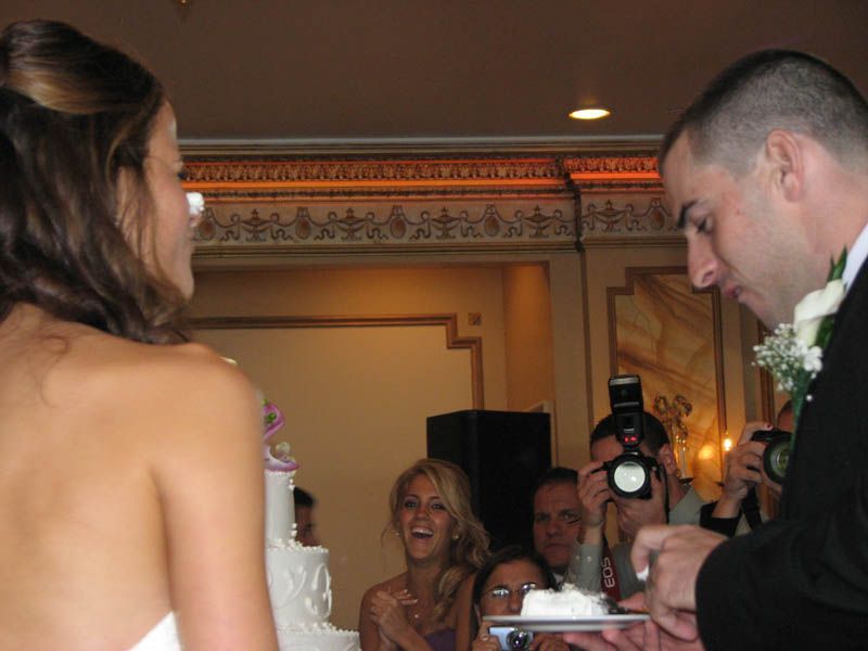 Bride and groom cutting wedding cake at reception, guests watching and photographing.