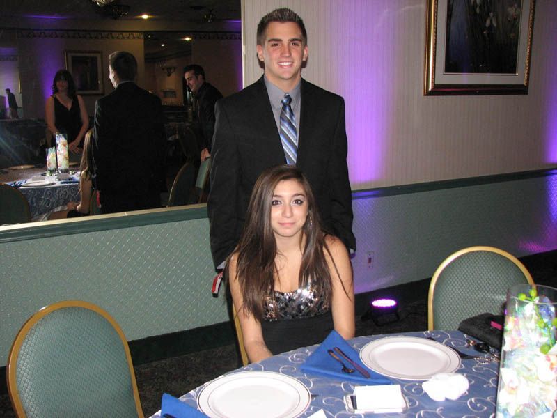 Woman seated at a table with a man standing behind her in a formal setting; purple lighting.