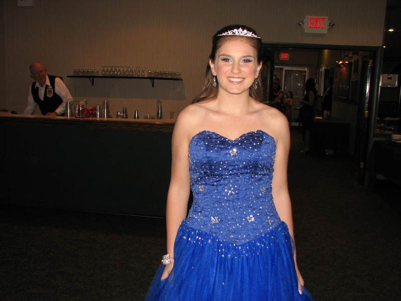 Woman in blue formal dress and tiara smiling, inside an event venue.