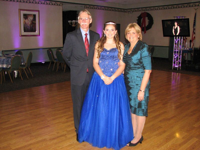 Family posing on a dance floor: teen in blue dress flanked by a man in a suit and a woman in a teal dress.