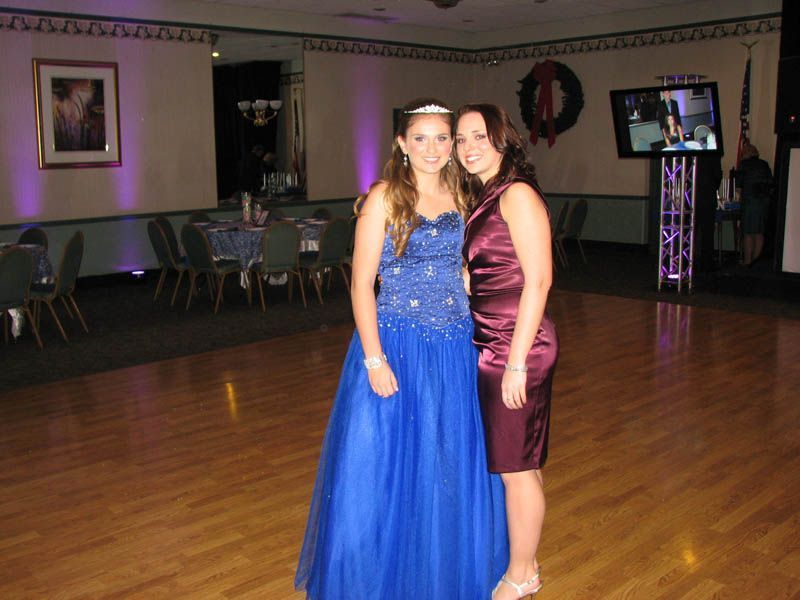 Two women posing at an event. One in a blue gown, the other in a burgundy dress, smiling. Wooden dance floor.