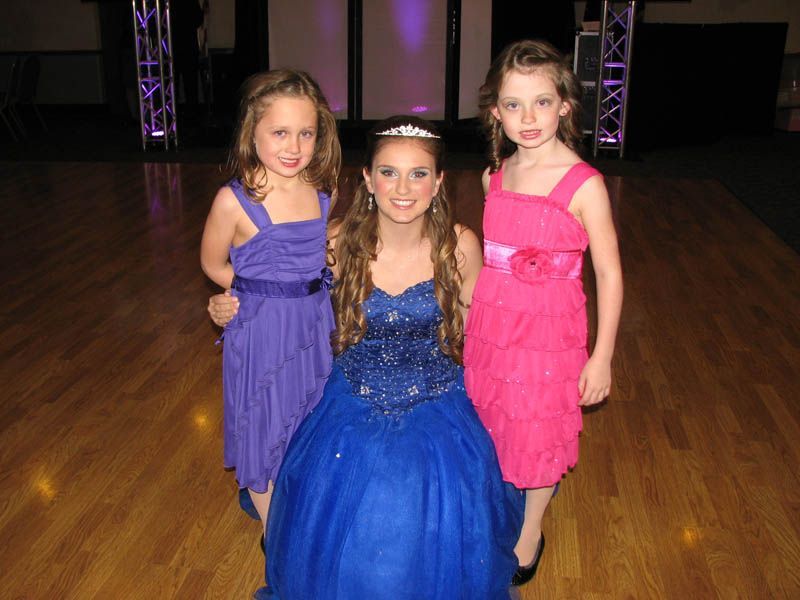 Three young girls smiling. One in blue gown with tiara, flanked by two in purple and pink dresses on a dance floor.