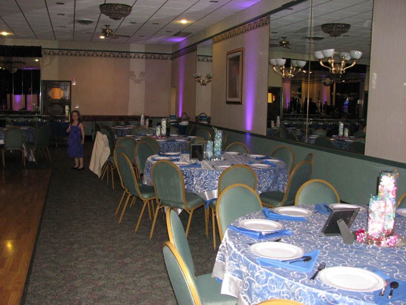 Reception hall with round tables set for a party, blue tablecloths, green chairs, a girl standing by a doorway.